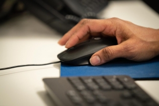 Close-up photograph of a person's hand using a computer mouse on a blue mouse pad. A keyboard is visible out of focus in the foreground, with a desk workspace in the background.