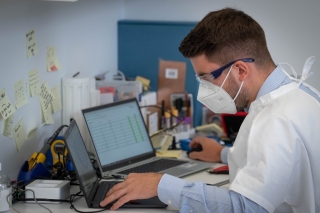 A person wearing a white protective gown, face mask and safety glasses working at a desk looking at two laptops. The person reviews data displayed on a laptop screen.