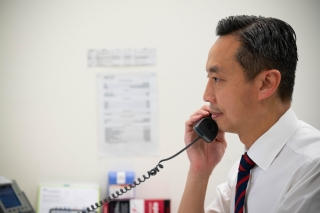A person in business attire holds a corded telephone handset to their ear while seated at a desk in an office. A desk phone and documents are visible in the background.