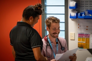 Healthcare staff review paperwork at a workstation in a clinical setting. One person seated with a stethoscope around their neck reads documents while another person stands beside them. Medical supplies and reference materials are visible on the wall, with a red door and window in the background.