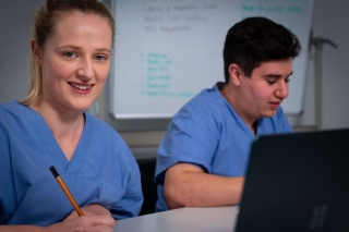 Two healthcare professionals in clinical uniforms working at a computer workstation. One person is writing notes while another uses a laptop. A whiteboard is visible in the background, illustrating a healthcare or clinical work environment.